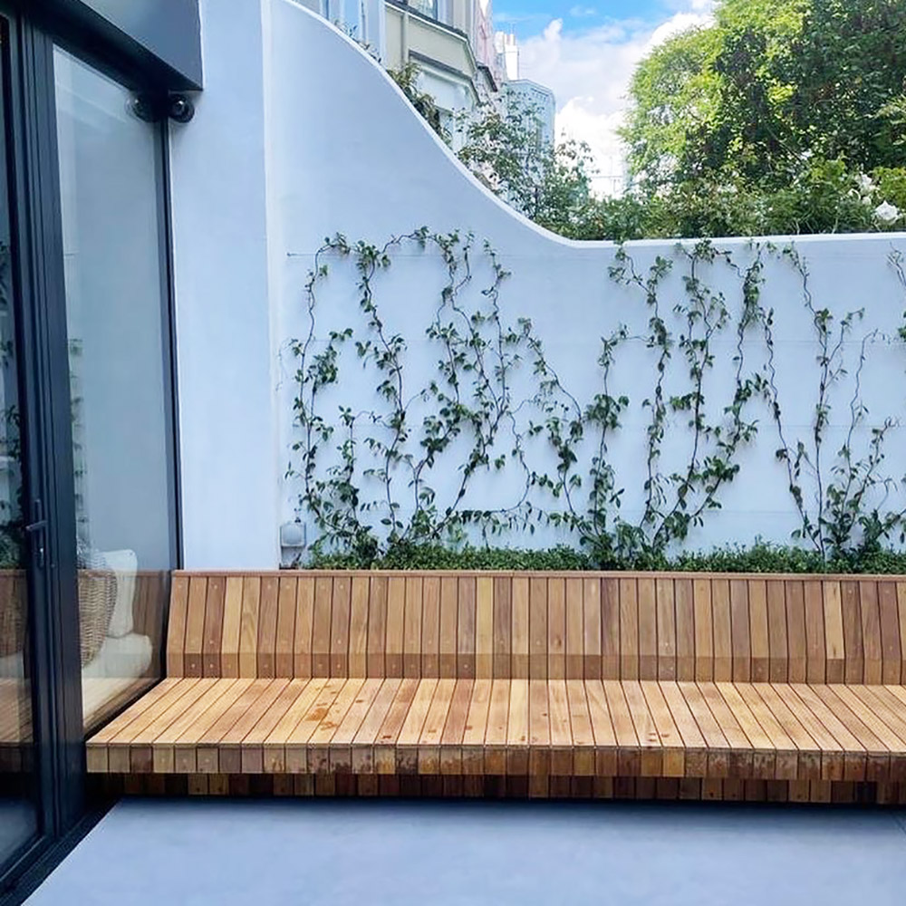 slat view of Iroko garden seat in a paved outdoor area with foliage growing up a white wall