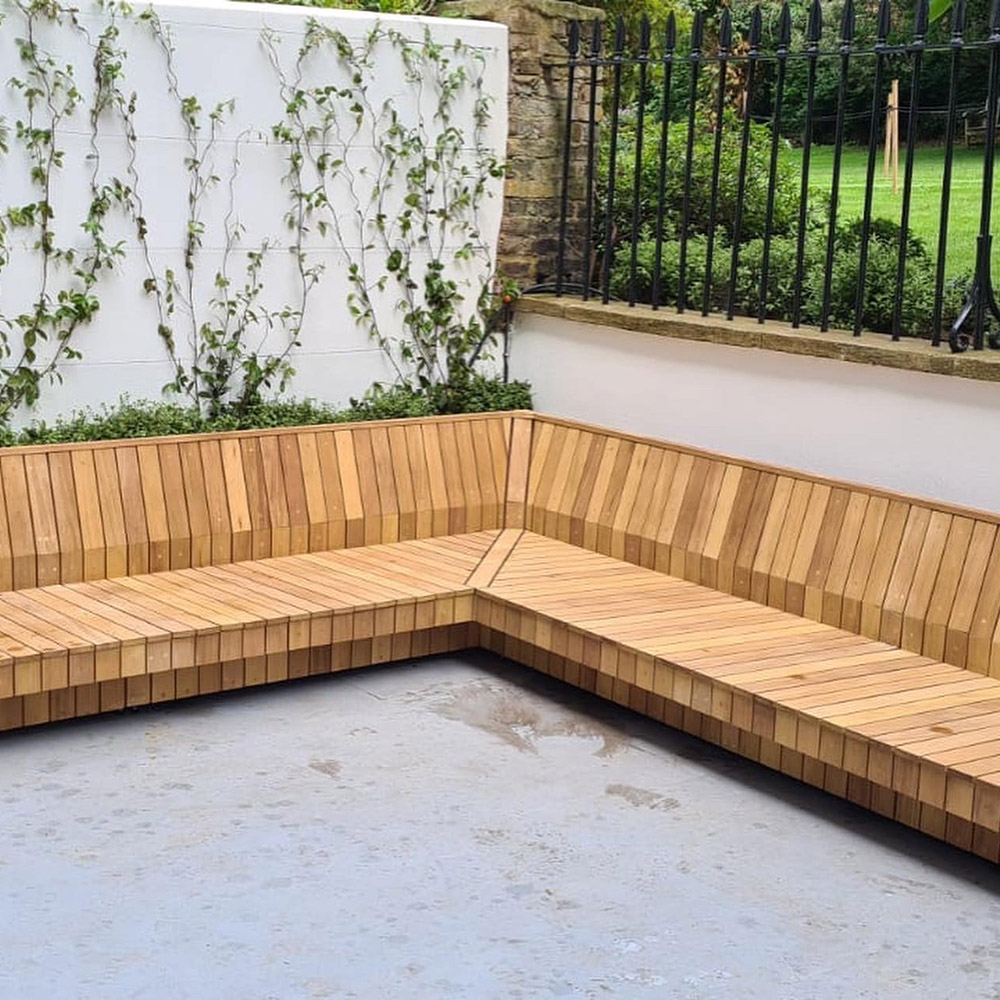 corner view of Iroko garden seat in a paved outdoor area with foliage growing up a white wall